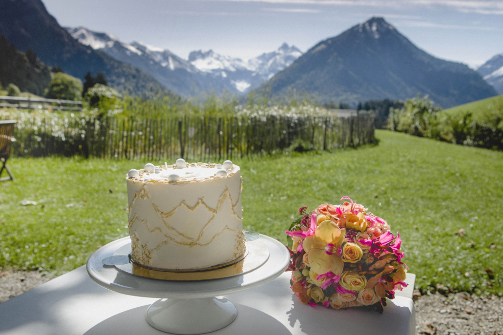 Hochzeitstorte mit Bergmotiv und Brautstrauß vor Alpenpanorama